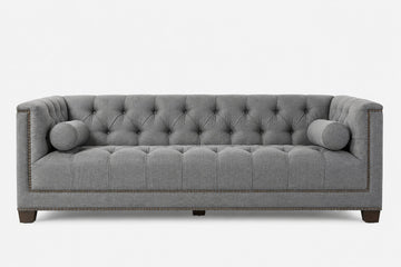 A gray tufted fabric sofa with cylindrical bolsters, nailhead trim, and dark wooden legs, shown against a plain white background.