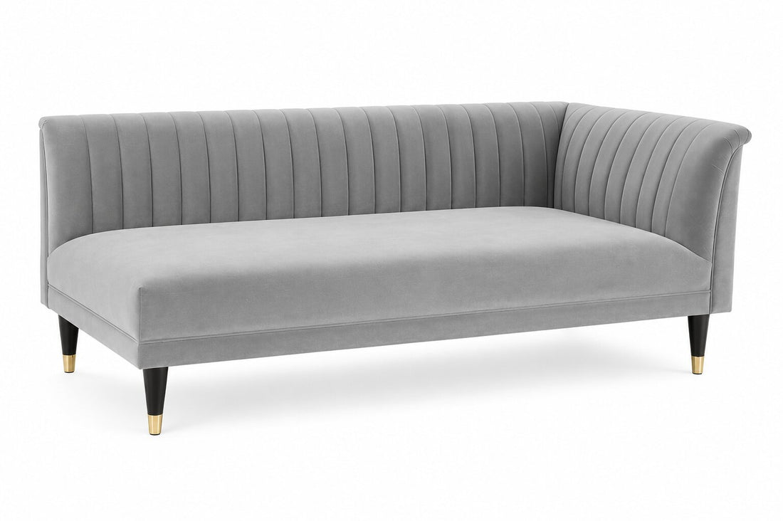 A modern gray velvet corner sofa with vertical channel tufting on the backrest and arms, supported by black tapered legs with gold caps, shown on a white background.