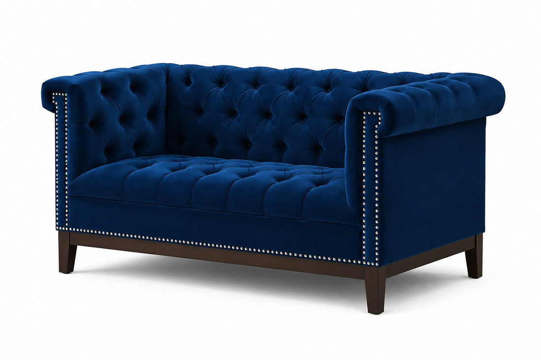 A classic Chesterfield loveseat sofa upholstered in deep navy blue velvet, featuring tufted button detailing, rolled arms, silver nailhead trim, and dark brown wooden legs, shown against a plain white background.
