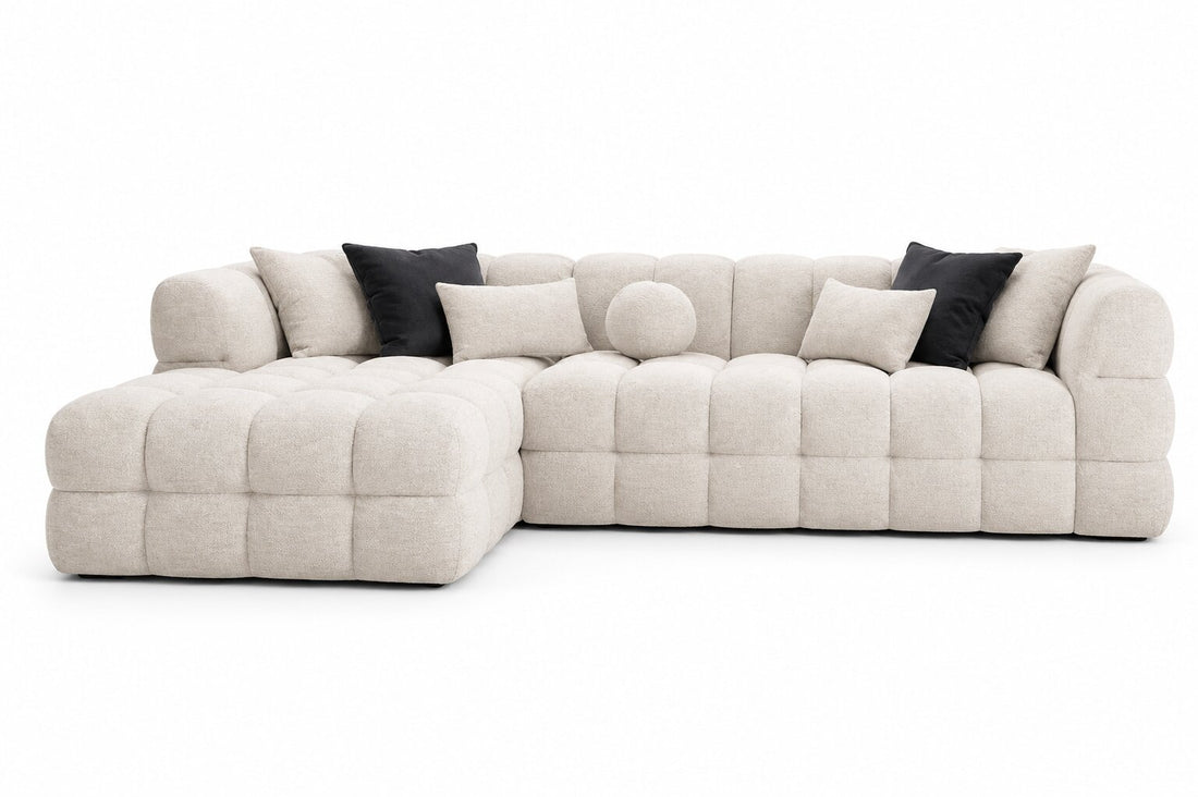 A modern modular sectional sofa in a light beige tufted fabric, styled with black accent pillows and a matching round throw pillow, set against a plain white background.