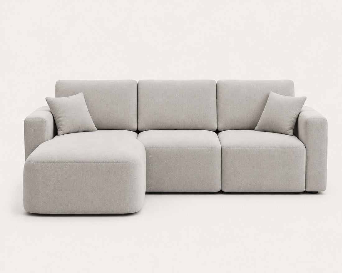 A modern modular sectional sofa upholstered in a light cream fabric, featuring a low-profile chaise lounge on the left, two back cushions, and a right armrest, shown on a solid white background.