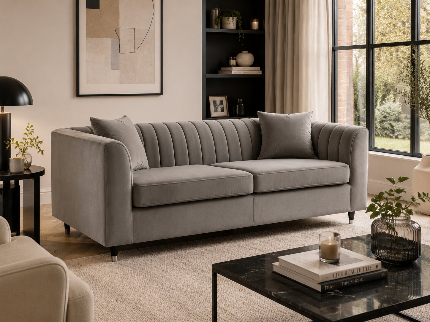 A light gray upholstered sofa with channel tufting on the back, shown in a styled living room setting with a black coffee table, side table, and large window.